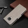 A gray leather-like flip case for a smartphone is shown on a textured black surface, with a brown notebook or wallet in the background and a pen to the left. Produktbild-Vorschau