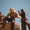 Three young women are sitting and standing on rocks outdoors, with a clear blue sky in the background. One woman is holding a pink object, possibly a speaker, and appears to be interacting with the other two. Produktbild-Vorschau
