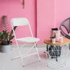 A white folding chair is shown in a room with pink walls and a patterned floor. A small table with drinks and a bag is next to the chair. Anteprima dell’immagine prodotto