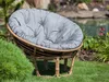 A round gray cushion on a rattan chair in a garden setting with a pond and plants in the background. Produktbild-Vorschau