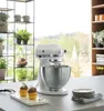 A white KitchenAid stand mixer sits on a kitchen counter next to two cupcakes, with shelves of plants and a window view in the background. Produktbild-Vorschau