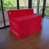 A red foldable mattress is shown in a room with large windows overlooking greenery. Náhled obrázku produktu