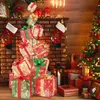 A festive Christmas scene with stacked LED gift boxes in front of a brick fireplace, stockings hanging, and a Christmas tree to the side. Produktbild-Vorschau