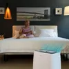 A woman sits on a bed in a bedroom, holding a remote control, with an air purifier operating in the foreground. Produktbild-Vorschau