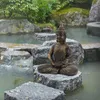 A bronze Buddha statue sits in a meditative pose on a rock in a tranquil garden pond with flowing water and lush greenery in the background. Náhľad obrázku produktu