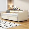 A beige upholstered daybed with a pull-out trundle, featuring a built-in shelf with LED lighting and decorative nailhead trim, set in a room with a lamp and framed pictures. Voir les images du produit