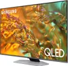 A Samsung QLED television is shown from a slight angle, displaying a colorful abstract image on its screen. The TV is placed on a light gray stand. Produktbild-Vorschau