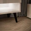 A close-up view of the legs and bottom shelf of a white TV stand with black legs, placed on a wooden floor. Náhled obrázku produktu