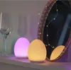 Two egg-shaped lamps, one pink and one yellow, sit on a shelf next to a wine glass and a decorative mirror with fairy lights. Produktbild-Vorschau
