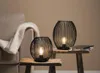 Two black wire lanterns with lit bulbs are placed on rustic wooden stools next to a sofa with a white blanket. Anteprima dell’immagine prodotto