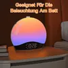A sunrise alarm clock with a gradient light display sits on a nightstand next to a stack of books. Produktbild-Vorschau
