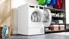 A white Bosch Serie 6 heat pump dryer with its door open, placed in a laundry room setting with a basket of clothes, shelves with folded towels and baskets, and colorful clothes hanging above. Produktbild-Vorschau