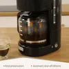 A close-up of a black filter coffee machine with a glass carafe filled with coffee. Steam is rising from a glass of coffee in the foreground. The background is a wooden surface and a light gray textured surface. Podgląd zdjęcia produktu