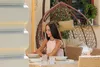 A woman sits at a table set for a meal on a patio, with a hanging chair in the background. Náhled obrázku produktu