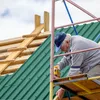 A construction worker is installing green corrugated metal roofing panels on a wooden roof structure, using a yellow power drill. Scaffolding is visible. Voir les images du produit