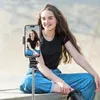 A young woman is sitting outdoors and taking a selfie with a selfie stick and phone. Anteprima dell’immagine prodotto