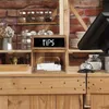 A wooden tip box with a chalkboard top labeled "TIPS" sits on a wooden counter in front of a brick wall. Other items like jars, a bell, and a cash register are visible in the background. Náhled obrázku produktu