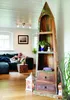 A living room scene with a person relaxing on a sofa and a unique boat-shaped bookshelf. Produktbild-Vorschau