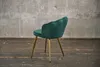 A green velvet chair with gold legs is shown against a concrete wall and wooden floor. Produktbild-Vorschau