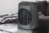 A gray electric heater is placed on a glass table in a room with a blurred background of a couch and blankets. Voir les images du produit