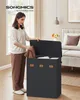 A woman is putting laundry into a three-compartment laundry basket in a bedroom. Produktbild-Vorschau