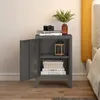 A gray metal nightstand with its door open, placed next to a bed. The nightstand has two shelves, with books and a decorative lantern on them. A camera and a vase with greenery are on top of the nightstand. Náhled obrázku produktu