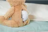 A close-up shot of a teddy bear sitting on a bed with a white sound module visible in a cut-out on its back. Anteprima dell’immagine prodotto
