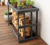 A dark gray wooden log rack filled with firewood sits on a wooden deck next to a white wall. Several potted plants are on top of the log rack. Podgląd zdjęcia produktu