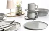 A collection of grey ceramic tableware, including plates, bowls, and cups, arranged on a white surface. A small lamp and a vase with flowers are also visible. Produktbild-Vorschau