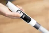 A hand is holding the handle of a white cleaning device, with a digital display showing battery life and mode. Voir les images du produit