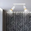 A white ceiling-mounted track light with two adjustable spotlights, each with a white cylindrical body and a gold accent at the end, illuminates a dark gray brick wall with a herringbone pattern. Curtains are visible on the left side. Podgląd zdjęcia produktu