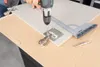 A person is using a drill to attach a hinge to a cabinet door, with a jig and ruler in place. Voir les images du produit