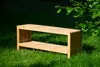A wooden bench with a firewood compartment sits on a grassy lawn, with green leaves from a tree overhead. Produktbild-Vorschau