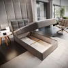 A bedroom scene with a bed frame open to reveal storage space, a side table with lamps, and a chair in the background. Produktbild-Vorschau
