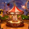 A miniature carousel with horses and LED lights, set against a blurred background of a fairground or amusement park. Produktbild-Vorschau