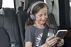 A young girl wearing headphones and looking at a smartphone while sitting in a car. Produktbild-Vorschau