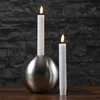 Two white LED candles are lit, one in a silver spherical holder and the other standing alone, against a dark stone wall. Produktbild-Vorschau