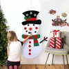 A young girl is decorating a large felt snowman with buttons, with a Christmas tree and wrapped gifts in the background. Náhľad obrázku produktu