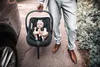 A person in a suit is holding a baby car seat with a baby inside next to a car. Produktbild-Vorschau