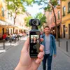 A hand holds a small digital camera, showing a selfie on its screen, with a blurred background of a street with buildings and people. Produktbild-Vorschau
