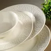 A close-up view of white porcelain dinnerware with embossed decorative patterns, stacked on top of each other. A small green plant is visible in the upper right corner. Produktbild-Vorschau