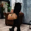 A person is holding a brown handbag while standing in a room with a leather armchair and plants. Podgląd zdjęcia produktu