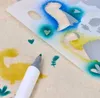 A close-up view of a stencil being used to airbrush a design onto fabric, with a marker pen visible. Produktbild-Vorschau