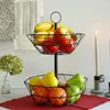 A two-tiered fruit basket filled with various fruits, placed on a table with decorative towels and candles in the background. Produktbild-Vorschau