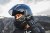 A person wearing a black motorcycle helmet and gloves, looking to the side with a mountain landscape in the background. Anteprima dell’immagine prodotto