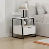 A modern white nightstand with black metal legs next to a bed. Produktbild-Vorschau