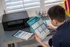 A young boy is sitting at a desk, looking at a printed exam week schedule and a tablet displaying the same schedule. A printer is visible on the desk. Podgląd zdjęcia produktu