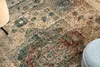 A close-up view of a vintage-style rug with intricate patterns in beige, cream, blue, and red tones. Parts of a wooden chair leg and a gray ottoman are visible at the edges. Voir les images du produit