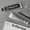 Two tubes of Marvis toothpaste are laid out on a gray surface next to a toothbrush and a glass of water, with a magazine in the foreground. Anteprima dell’immagine prodotto