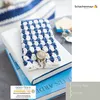 A crocheted blue and white bag with a button closure rests on top of two books. A logo and certification mark are visible. Produktbild-Vorschau